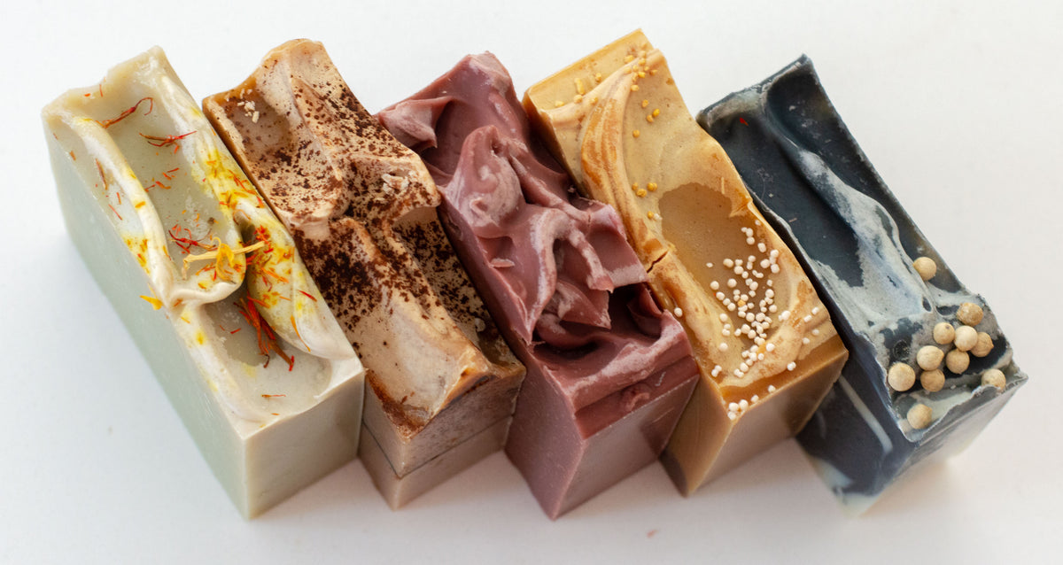 Six artisanal handmade soap bars arranged in a row on a white background, featuring various colors and textures including marbled, swirled, and speckled designs.